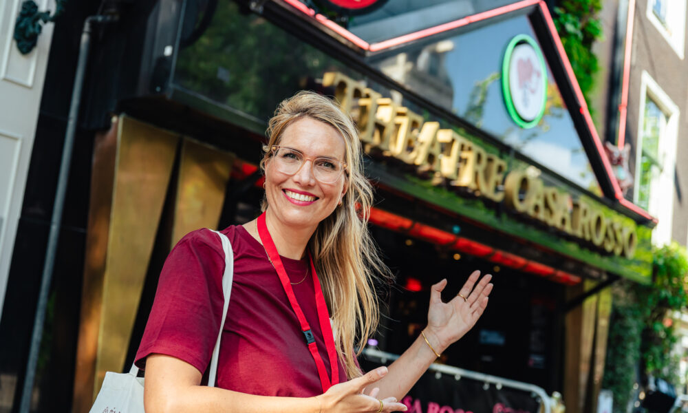A smiling woman standing outside a cafe in Amsterdam. She is wearing a red shirt and a lanyard.