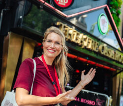 A smiling woman standing outside a cafe in Amsterdam. She is wearing a red shirt and a lanyard.