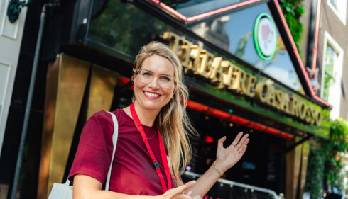 A smiling woman standing outside a cafe in Amsterdam. She is wearing a red shirt and a lanyard.