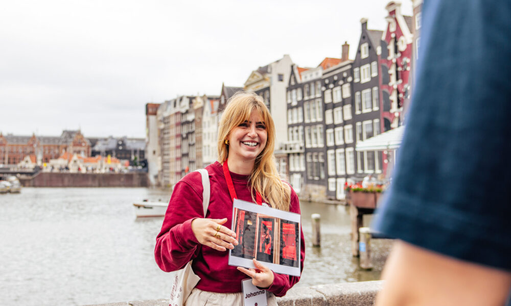 A smiling woman holding a city guide in Amsterdam. She is standing by a canal with traditional Dutch buildings in the background.