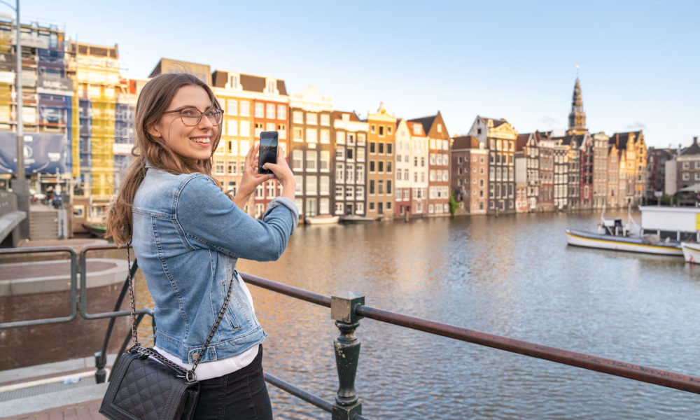 Woman taking a photo with a smartphone by a river. A cityscape with buildings and a boat is in the background.
