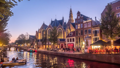 A serene Amsterdam canal at dusk with boats and buildings. People enjoy the evening.