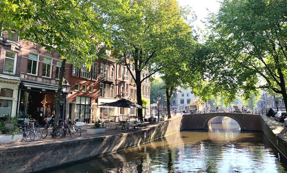 A serene canal scene in Amsterdam with a small bridge. The waterway is lined with trees and historic buildings.