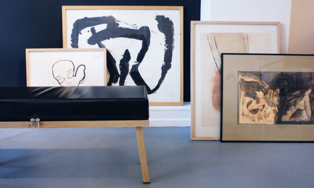 Modern art pieces on display in a room. A black bench with wooden legs in front.