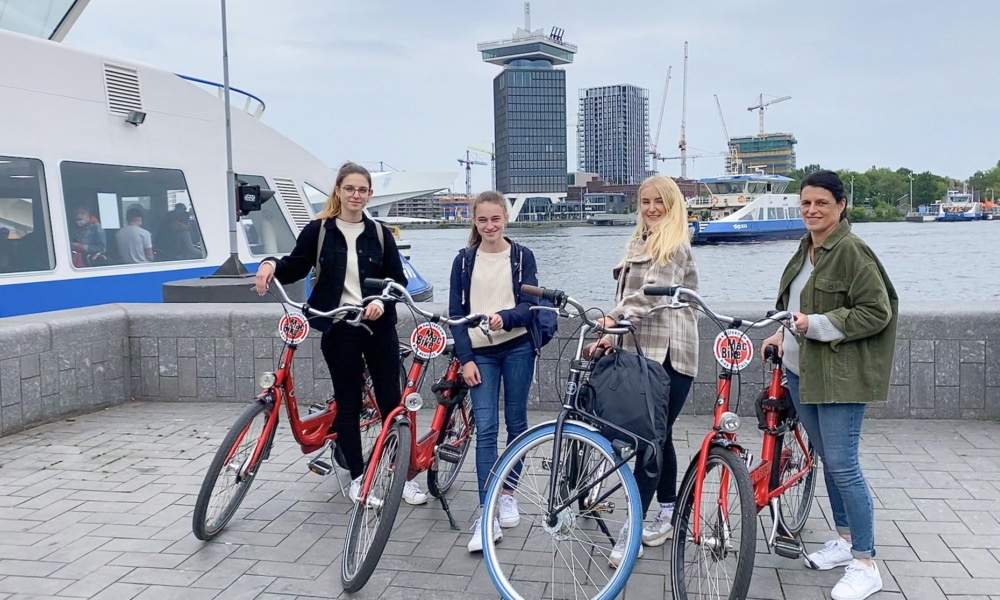 Four women standing with bicycles by the water. They are smiling and looking at the camera.