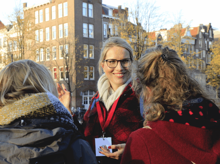 Blonde woman wearing a red jacket and lanyard talking to tourists. City buildings in the background.