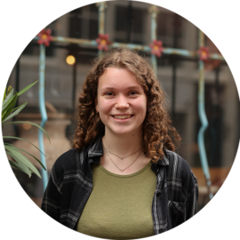 A young woman with curly hair smiling. She is wearing a green shirt and a black plaid shirt.