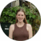 A smiling woman with long brown hair, wearing a brown tank top and a necklace. She is standing in front of green plants.