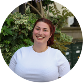 A smiling woman with red hair standing in front of lush greenery. She is wearing a white shirt.