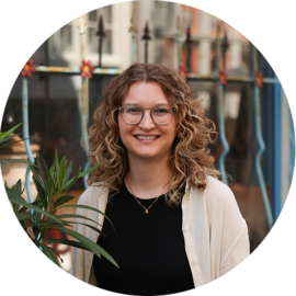 A woman with curly hair and glasses smiling at the camera. She is wearing a black top and a beige cardigan.