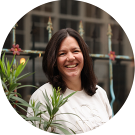 A smiling woman with dark hair standing in front of green plants. She appears to be on a city trip in Amsterdam.
