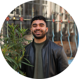 A man with a warm smile wearing a black leather jacket. He is standing in front of a plant and a metal fence.
