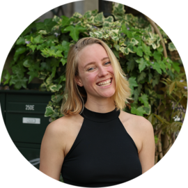 A smiling woman in a black top standing in front of greenery. She appears to be on a city trip in Amsterdam.