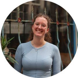 A young woman with brown hair wearing a blue top smiling at the camera. She appears to be outdoors.