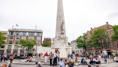 Dam Square with Obelisk and people sitting on steps. The National Monument in Amsterdam.